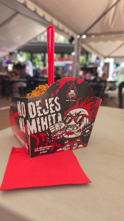 A close-up, eye-level shot focuses on a red plastic utensil sticking out of a partially filled, black and red cardboard food container. The container features bold white text that reads "NO DEJES NI MIHITA" across its front and a stylized graphic of a skull wearing sunglasses and holding a burger. To the right of the text, the brand logo "NI MIHITA" is visible in red and white. Below the graphic, an address is printed in white: "C/ DR. JAIME GARCIA ROYO 16 18014, GRANADA - SPAIN". A folded red napkin sits in front of the container. The background is blurred, showing a tented outdoor seating area with people and tables, suggesting a food stall or casual dining establishment, possibly in the city of Almuñécar, Spain, given the address format and the nature of the establishment.