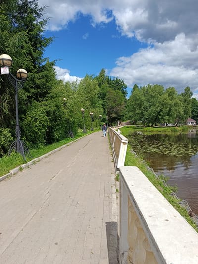 A paved walkway lined with trees and decorative lampposts curves alongside a body of water. A lone individual walks away from the camera on the path, enjoying a stroll. The water is dotted with lily pads, and a building with a white roof and dark beams is visible in the distance across the water. The sky is a vibrant blue with scattered white and gray clouds, suggesting a pleasant, partly cloudy day. The scene evokes a sense of tranquility and leisure in a park-like setting. A sign on one of the lampposts displays text in Russian, which reads "ВЪЕЗД" and "ЗАПРЕЩЕНО" followed by "ДЛЯ ПОСЕТИТЕЛЕЙ" indicating "ENTRY PROHIBITED FOR VISITORS".