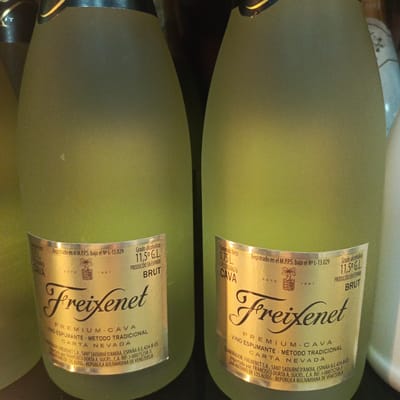 Two bottles of Freixenet Brut cava are visible in the foreground, standing next to each other. The bottles are a pale green color with a frosted finish. Both bottles have prominent labels with the Freixenet logo in black script. The labels contain information in Spanish, including "PREMIUM CAVA," "VINO ESPUMANTE," "METODO TRADICIONAL," and "CARTA NEVADA." They also state the alcohol content as "11,5º G.L." and that the product is "PRODUCIDO EN ESPAÑA." One bottle's label indicates "Contenido Neto 0,75 L." and the other mentions "ESTD 1861." Below this text, one label reads "ELABORADOR: FREIXENET S.A., SANT SADURNÍ D'ANOIA, ESPAÑA R.E.424-B-ES" and "IMPORTADO POR: FRANCISCO DORCA A. SUCRS., C.A. RIF: J-00075258-3 ANTONIO DE LOS ALTOS - REPÚBLICA BOLIVARIANA DE VENEZUELA." This text places the context of importation and distribution within Venezuela, specifically mentioning "ANTONIO DE LOS ALTOS" which is a sector of the city of Los Teques. The background is dark and out of focus, suggesting an indoor setting, possibly a store or a cellar. The lighting is somewhat dim, with a yellowish hue. No people are visible in the image.