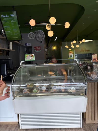 The image depicts the interior of "Shake Me Gelateria," a dessert shop located in AMCO, Area Metropolitana Centro Occidente, Colombia. The scene is characterized by a vibrant green ceiling and upper walls, complemented by light wood paneling and flooring.
In the foreground, a curved glass display freezer showcases various gelato flavors, each labeled with a small cone-shaped marker. To the left, a person's hand holds a white cup with a wooden spoon, suggesting a recently served or purchased item.
Behind the counter and the freezer, a female employee is partially visible, wearing a light-colored shirt and a green patterned apron. She is actively reaching into an area behind the main display, possibly preparing an order or attending to items. Above the counter, a black monitor for a point-of-sale system is visible.
The environment includes two modern Sputnik-style pendant light fixtures with multiple spherical globes illuminating the space. A large digital menu board on the upper left displays "Shake Me Gelateria" and various categories such as "Gelato," "Cafe," "Postres," "Bebidas Frias," and "Extras," with prices listed in Colombian Pesos. Two wall-mounted fans are visible on the grey wall section. Signage includes "COCINA / KITCHEN," a red "Coffee" sign, and a "New York Cookies / DISFRÚTALAS AHORA!" promotional sign. A promotional image for a dessert cup is also displayed to the right of the main counter area.