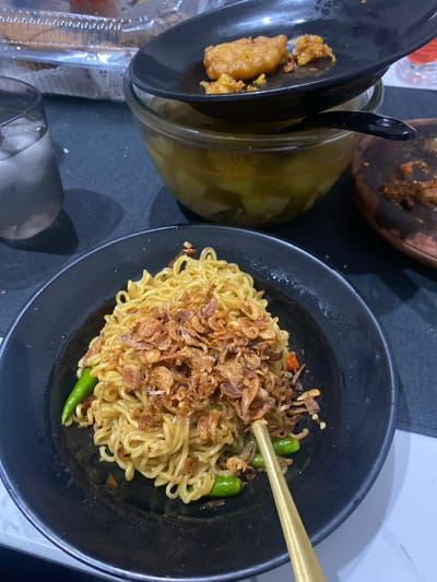 A close-up, eye-level, full shot captures a bowl of instant noodles, topped with fried shallots and garnished with green chilies, positioned on a dark tablecloth. A golden fork rests inside the bowl. In the background, a glass filled with ice water is visible, along with a clear plastic container holding a cake and a glass bowl containing a yellow soup with pieces of food. A black plate with fried food sits atop the soup bowl, and a wooden mortar with chili residue is to the right. The scene appears to be a casual dining setting, likely within a residential or small eatery context in Cibinong, Indonesia, given the presence of common Indonesian food items. The lighting suggests it may be nighttime or an indoor setting with artificial light.