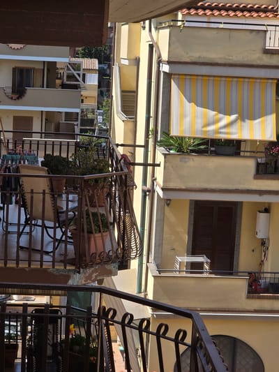 The image captures a view of apartment balconies in Ladispoli, Italy. The balconies are constructed of metal railings and are adorned with potted plants and outdoor furniture. On one balcony, a striped awning in yellow and white is extended. Another balcony features a clothes drying rack. The buildings are painted in warm, sun-baked tones of yellow and cream, with terracotta roof tiles visible on the top structures. Sunlight casts shadows across the scene, indicating a bright, clear day. There are no people visible in the image.