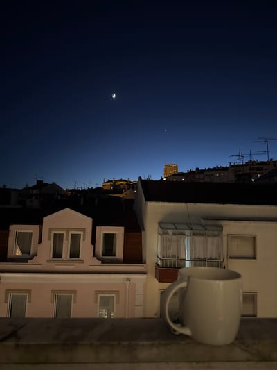 The image depicts a nighttime view of urban rooftops in Algés, Portugal, with a crescent moon and a bright celestial object visible in the deep blue sky. In the foreground, a white mug sits on a ledge, partially obscuring the lower portion of a building with pink and white facades. Several windows are visible on these buildings, some illuminated. Beyond the immediate buildings, a cluster of structures with antennas and chimneys stretches across the horizon, with a taller, illuminated building in the distance. The overall scene conveys a quiet, twilight atmosphere overlooking a residential area.