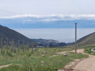 The image captures a scenic vista in Cholpon Ata, Kyrgyzstan, with a focus on the landscape and distant bodies of water. In the foreground, a dirt path winds through a grassy area dotted with rocks. A wire fence, constructed with concrete posts and wire, runs along the left side of the path. In the mid-ground, a utility pole stands prominently, supporting several wires that stretch across the frame. Beyond the pole, a body of water, likely Lake Issyk-Kul, stretches towards the horizon, characterized by its calm surface and the faint outlines of distant clouds. A town or village is visible along the shoreline, with buildings and trees forming a settlement. The background is dominated by a vast, pale sky with scattered clouds. The overall impression is one of a natural landscape with subtle signs of human presence.