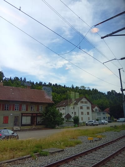 The image depicts a roadside scene with buildings and a forested hill in the background. In the foreground, a train track runs diagonally across the frame. To the left, a dark colored car is parked on the side of the road in front of a building with a red-shuttered window and several other windows. The building appears to be a storefront with a large glass window. Further down the road, two white cars are parked in front of a larger, light-colored building with multiple windows. A white van is visible in the distance to the right. The environment appears to be a semi-rural or suburban area with a clear sky and clouds. The location is identified as Riken, Switzerland.