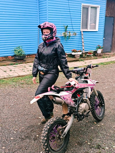 In front of a bright blue corrugated metal building in Rassol'naya, Russia, a person stands beside a dirt bike. The person, wearing a pink motocross helmet, black jacket, and black pants, has a fanny pack around their waist. The dirt bike is white and pink with purple rims. The ground is covered in gravel and scattered leaves. The building has a white-framed window and a dark gray door. Several potted plants are visible around the building.