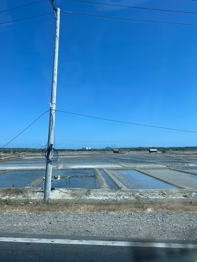 The image depicts a salt field under a clear blue sky in Jeneponto, Indonesia. In the foreground, a grey utility pole stands with coiled black wires and cables attached to it. The pole is positioned on a rough, gravelly surface next to a paved road.
The main focus of the scene is the expansive salt field, which is divided into numerous rectangular ponds filled with shallow water. The ponds appear to be in various stages of salt production, with some reflecting the sky and others showing a lighter, more solid surface, likely accumulated salt. The edges of the ponds are lined with dirt and dry vegetation.
In the middle ground, several small, dark-roofed structures are scattered across the landscape. Further in the distance, a power plant with two tall smokestacks can be seen against the horizon. The surrounding terrain is relatively flat, with a line of trees and vegetation in the far background. No people are visible in the image. The overall impression is one of an industrial or agricultural landscape dedicated to salt production.