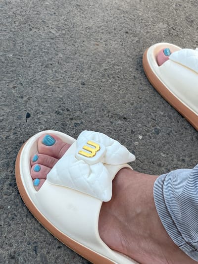 A close-up, eye-level shot shows a person's feet wearing white slides with a decorative puffy bow that has a gold "3" emblem. The slides have tan soles and are resting on a concrete surface. The toes are painted with bright turquoise nail polish. The person is wearing light-colored, vertically striped pants on their right leg. The scene is set outdoors on a paved surface, possibly a sidewalk or road, in Palanca, Moldova. The lighting suggests it is daytime, with the sun casting some shadows.