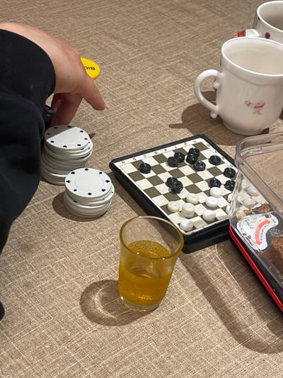 The scene depicts an indoor setting in Jisr az-Zarqa, Israel, featuring a table laden with gaming components, beverages, and a snack. A human left hand, with manicured nails, is visible in the upper left quadrant, positioned above two stacks of white poker chips adorned with blue spade symbols. The larger stack comprises approximately twenty chips, while the smaller contains about ten. A small, yellow circular disc marked "BIC" rests near the hand.
Centrally located is a small, portable checkers board with black and white squares, featuring algebraic notation "a, b, c, d" along one edge. Numerous black and white checkers pieces are distributed across the board, suggesting an ongoing or recently concluded game. To the right of the board, a clear glass contains an amber-colored, effervescent liquid. In the upper right, two white ceramic mugs are present; one features a small red graphic, and the other, partially visible behind it, displays red polka dots. A clear plastic container with a red base, containing remnants of a baked good or crumbs, is positioned on the far right. All items rest on a light brown, textured surface, possibly a woven tablecloth, under directional lighting that casts shadows.