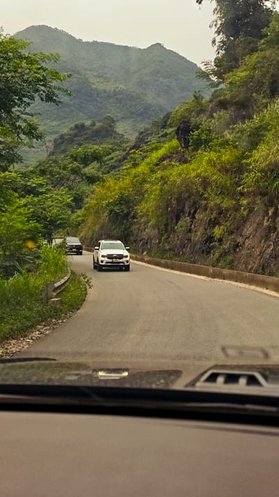 The image depicts a winding road in the Cán Tỷ Commune, Vietnam, as viewed from inside a moving vehicle. Two cars are visible ahead on the road: a white SUV in the immediate foreground and a dark-colored car further behind. Lush, green vegetation covers the surrounding hills and mountains. The road is asphalt and has concrete barriers on one side. The sky is overcast.
