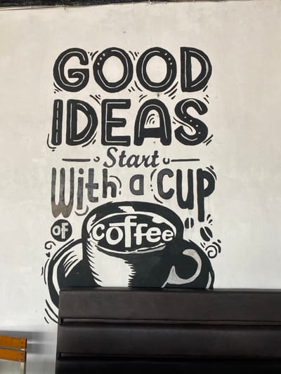 This image depicts an indoor scene in Aluebutuha, Indonesia, focusing on a white wall adorned with a black hand-drawn mural. The mural features the phrase "GOOD IDEAS Start With a cup of coffee" in decorative block and script lettering, accompanied by an illustration of a large coffee cup with the word "coffee" inside. Stylized swirls and small coffee bean motifs accent the design. Below the mural, a dark brown, multi-paneled upholstered bench or booth seating is positioned, obscuring the lower portion of the coffee cup illustration. To the left of the bench, a section of a wooden chair with a silver metal frame is partially visible. The wall surface shows minor imperfections and faint smudges. The upper edge of the wall, where it meets the ceiling, is visible at the top of the frame. No individuals are present in the scene.