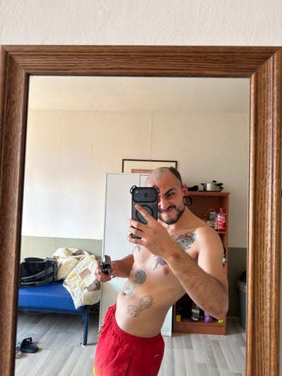 The media file is a mirror selfie of a person. The subject is a shirtless person with tattoos, wearing red shorts, holding a smartphone in their hand, and looking at the camera. The person has dark hair, a beard, and is wearing large gauges in their ears. In the reflection, we can see a shelf unit with various items, including bottles and other objects, a trash can, and a bag. The setting appears to be indoors, likely a room in a home, with wooden flooring visible in the reflection. The lighting suggests it is daytime. There is no visible text in the image or its reflection, except for potential branding on the bottles in the background, which are not clearly legible. The overall tone of the image is casual and personal, typical of a mirror selfie. The location context of Boxtel, Netherlands, is provided separately and is not visually discernible from the image itself.