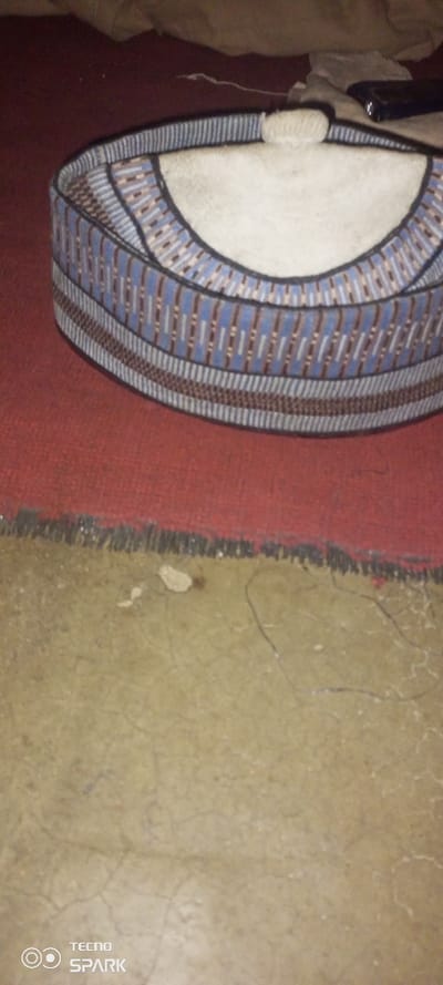 A traditional Hausa Fulani cap, known as a 'Fula cap,' rests atop a red carpet. The cap is cylindrical with a flat, round top. It features intricate embroidered patterns in shades of blue, white, and brown. The surrounding environment includes a tan-colored floor with visible cracks and a blurry backdrop of what appears to be a bed. A black object, potentially a phone, is vaguely visible near the top of the frame. The "TECNO SPARK" watermark is located in the bottom left corner.