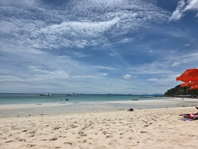 This media file depicts a tropical beach scene in Bang Sare, Thailand. The foreground is dominated by a sandy beach with footprints and small pebbles scattered across it. In the mid-ground, the turquoise water gently laps at the shore, revealing a shallow sandbar. Several inflatable mats are visible in the shallow water, suggesting leisure activities.
Further out at sea, there are several boats, including two larger vessels with slides, indicating they are likely tourist boats or ferries. In the distance, a lush green, hilly island forms the backdrop to the right of the frame, with a few people walking along the shoreline.
To the far right of the image, a bright orange umbrella is partially visible, casting a shadow on the sand, with a person lying down underneath it, perhaps relaxing or sunbathing. A life jacket and other personal items are also visible near the umbrella.
The sky is a vibrant blue, dotted with fluffy white clouds that create a dynamic pattern. The overall impression is one of a bright, sunny day, likely late morning or early afternoon, given the sunlight and cloud formations. The scene evokes a sense of tranquility and leisure, characteristic of a tropical vacation destination. There is no visible text in the image.