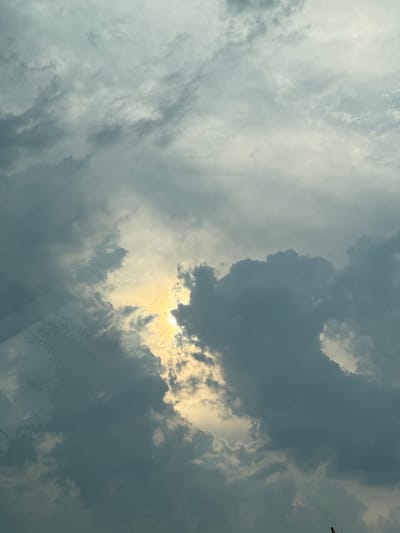The image captures a dramatic sky filled with an array of clouds. The predominant subjects are the clouds themselves, which vary in density and color, ranging from light, wispy formations to dark, imposing masses. The sun is partially obscured by these clouds, casting a bright, golden light through a gap in the darker formations. This creates a striking contrast between light and shadow, highlighting the textures and shapes of the clouds.
The activity taking place is the interplay of light and shadow in the sky, indicative of changing weather conditions. The scene is an outdoor one, presumably taken from ground level looking upwards. The time of day is suggested by the sunlight, which appears to be either late afternoon or early morning, given its low angle and warm hue. The weather appears to be mixed, with signs of both bright sunshine and impending rain or a storm due to the dark clouds. There are no specific location cues visible in the image itself, apart from the general impression of an open sky.
Notable details include the dynamic patterns formed by the clouds and the way the light pierces through them. There are no people or man-made objects prominently featured, save for a very faint, dark vertical element in the top right corner which could potentially be part of a distant structure or a thin branch. The overall mood conveyed by the image is one of awe and perhaps a touch of foreboding, due to the dramatic and somewhat turbulent nature of the sky. There is no visible text in the image.