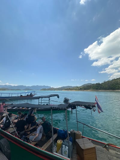 The image shows a longtail boat filled with people on Cheow Lan Lake in Ban Ta Khun, Thailand. The boat is oriented along the longitudinal axis with several passengers seated and standing. On the port side of the boat, the shoreline is visible, featuring mountains and lush greenery. The water of the lake is a greenish-blue color. The Thai flag is attached to the rear of the boat, alongside the boat's engine. The sky is blue with scattered white clouds.