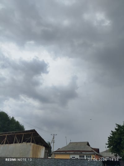 A medium shot captures a cloudy sky over a residential area in Асан-Чек, Kyrgyzstan. The sky is filled with heavy, gray clouds, suggesting overcast conditions. In the foreground, the roofs of several houses are visible. One building, on the left, has a light-colored facade and a weathered, corrugated roof supported by wooden beams. To its right, another house features a gray, tiled roof and a yellow facade with white-framed windows. A utility pole with wires stands between the houses. Trees are visible on the left and right edges of the frame. A black object, possibly a bird, is seen in the sky above the houses. The bottom left corner displays "REDMI NOTE 13" in white text, and the bottom right corner shows the date and time "21.06.2026 14:35" in white text.
