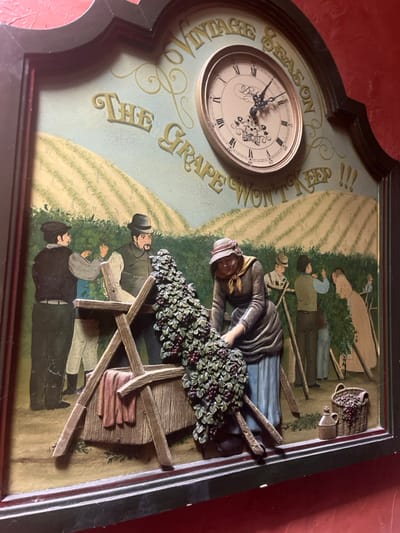 A framed wall decor piece, likely a clock, depicts a detailed vineyard harvest scene. The piece features a dark brown wooden frame with an ornate, arched top border. A circular clock with Roman numerals, black hands, and decorative central motifs is embedded in the upper right, indicating approximately 10:20. Above the clock, "VINTAGE SEASON" is written in raised yellow letters; below it, "THE GRAPE WON'T KEEP !!!" is similarly rendered.

The main scene below is a combination of painting and three-dimensional relief. It portrays a vineyard landscape with rolling hills of grapevines under a light blue sky. Six human figures, dressed in traditional attire, are engaged in grape harvesting and processing. On the left, three men are harvesting and interacting with a wooden grape-processing structure. One man in a dark hat and vest observes; another in a light shirt picks grapes; a third man in a fedora and vest stands by the wooden structure.

A prominent female figure, rendered in three-dimensional relief, is positioned centrally. She wears a brown headscarf, brown top, and blue skirt, leaning forward to gather a large, realistic cluster of dark grapes and green leaves (also in relief) into the wooden processing structure, which contains a reddish cloth. In the background on the right, two additional figures, a man and a woman, are visible harvesting grapes among the vines. In the lower right foreground, a wicker basket filled with dark grapes and a ceramic jug are present in relief. The entire piece is mounted on a reddish-orange wall within the city of Sélestat, France.