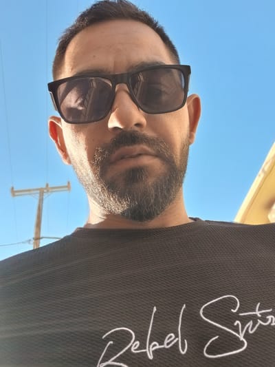 A close-up, low-angle outdoor selfie shows a man with dark hair, a beard, and sunglasses against a clear blue sky. The man is wearing a dark t-shirt with white embroidered text. The background features a utility pole with power lines. A building facade, likely painted yellow, is partially visible on the right side of the frame. The image appears to have been taken during daylight, with sunlight casting shadows on the man's face and clothing. The specific location in Malia Municipal Unit, Greece, is not discernible from the visual cues in the image.