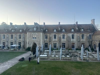 The image displays the exterior of a multi-story, historic-style stone building and its expansive courtyard, located in Cernay-la-Ville, France. The building features light grey stone masonry, a red-tiled roof with numerous dormer windows, and multiple brick chimneys. A large arched entryway, decorated with a conical Christmas tree, is centrally positioned on the facade, flanked by multiple rectangular windows with grey frames.
The foreground and midground comprise a green grass lawn, traversed by a diagonal stone pathway. Directly in front of the building, on either side of the pathway, are two distinct patio areas. These patios are furnished with numerous circular tables and chairs, predominantly covered in white protective shrouds. Several tall, slender, light-colored covered objects, consistent with outdoor heaters or retracted parasols, are distributed across both patio areas. A circular stone fountain is visible within the right-side patio section.
Two individuals are present on the grass lawn. One subject, dressed in dark clothing, is walking toward the left side of the frame. The second subject, wearing a long, light-colored overcoat, stands stationary, facing the building. The scene is illuminated by diffused natural light under a pale blue sky, indicative of early morning or late afternoon conditions.