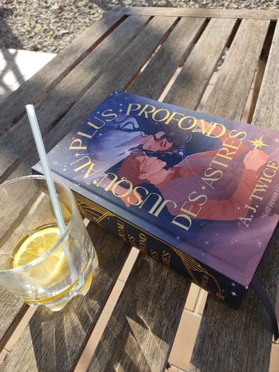 The scene depicts a static, slightly elevated, close-up view of objects resting on a weathered wooden slatted table in an outdoor setting in Aubagne, France. A hardcover book, approximately 24 cm by 16 cm, is positioned horizontally on the table. Its dark blue cover features a starry motif and gold text reading "AU PLUS PROFOND JUSQU'AU DES ASTRES" by "A.J. TWICE" (Gulf stream éd.). The cover artwork illustrates two stylized human figures, one with dark hair and one with reddish-brown hair and glasses, set against a cosmic background. A black fabric bookmark ribbon extends from the lower edge of the book. Adjacent to the book, on the left, is a clear glass, approximately 10 cm tall, with a ribbed lower section. It contains a transparent liquid, two yellow lemon slices, and a white straw with thin blue horizontal stripes. Direct sunlight illuminates the scene from the upper left, casting distinct parallel shadows from the table slats across the wooden surface and from the objects themselves. No individuals are present in the frame.