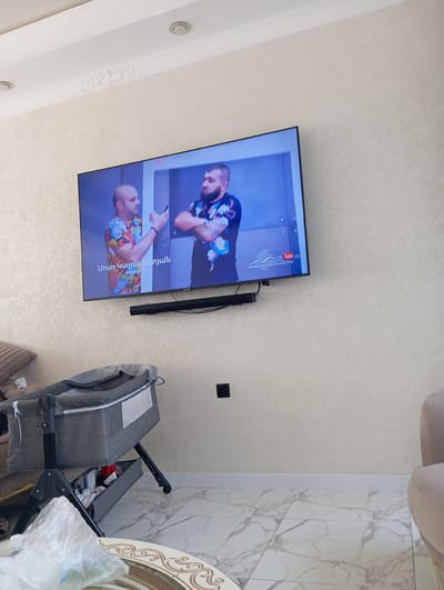 The image shows an indoor scene with a large television mounted on a textured wall. On the TV screen, two men are visible, with one holding a smartphone. Below the television, a black soundbar is mounted. To the left of the television, a grey baby crib with a canopy is positioned, and a stack of items is visible in the lower compartment of the crib. A plastic bag rests on a decorative table in the foreground. The floor is covered with marble-patterned tiles. A black electrical outlet is visible on the wall to the right of the crib. The room appears to be a living area or bedroom. The specific location in Nizhnyaya Saniba, Russia, is not visually identifiable from the image itself.