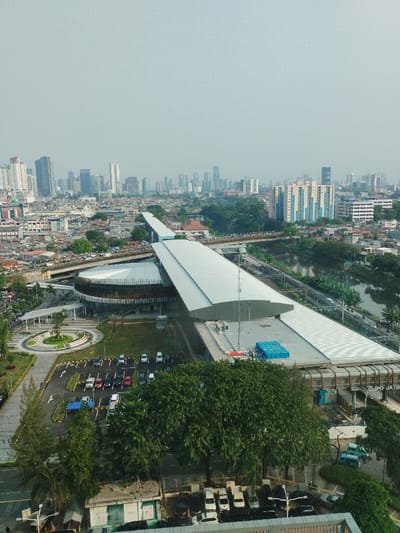 This high-angle, panoramic image depicts the Jatinegara LRT Station complex and its surrounding urban environment in the Special Capital Region of Jakarta, Indonesia. The central focus is a modern, elevated Light Rail Transit (LRT) station with a long, light-grey, curved roof, extending horizontally across the mid-ground. Adjacent to the elevated platform structure is a prominent, multi-story, circular building with a white, angled roof design and a dark, possibly wood-textured, facade. A large, paved parking lot occupied by approximately 20-30 passenger vehicles is situated in the immediate foreground, to the left of the circular building, surrounded by mature green trees and some manicured green spaces.
To the right of the elevated LRT station, a murky river or canal runs parallel, bordered by vegetation and smaller structures. Elevated roadways and bridges with visible vehicle traffic traverse the scene at various points. The urban landscape surrounding the station consists of densely packed low-rise residential and commercial buildings, interspersed with additional patches of greenery. In the distant background, a sprawling skyline of numerous high-rise commercial and residential buildings is visible under a hazy, pale sky, characteristic of a large metropolitan area. A blue tarp or covering is visible on the roof of the elevated station structure, and a communications tower protrudes from the same roof section. The text "JPO JATIBE" is faintly visible on the underside of the elevated station structure, confirming its location near a pedestrian bridge in the Jatinegara area. No individual people or specific actions are discernible due to the elevated vantage point.