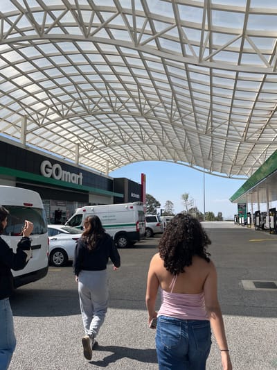 The image captures an outdoor scene at what appears to be a gas station and convenience store complex during daylight hours under a clear sky.

In the foreground, three individuals are walking away from the camera towards the right. The person closest to the viewer in the center has long, dark curly hair, wears a light pink strapless top, and blue jeans. To their left, another person with long dark hair is dressed in a black long-sleeved top and grey sweatpants. A third person, partially visible in the bottom right corner, wears a dark top with a white star design. Distinct shadows are cast on the asphalt ground, indicating bright sunshine.

To the left, a prominent architectural feature is a large, translucent white canopy with a grid-like metal frame, providing shade over the area. Below this canopy, a dark-colored building houses a "GO mart" convenience store, with its name visible in white text on a green and black sign. Above the "GO mart" sign, a red "McDonald's" sign with the golden arches is prominently displayed.

Several vehicles are present, including a white ambulance van with "AMBULANCIA" and "EMERGENCIA" visible on its side, and other white cars and vans parked in front of the building. In the background to the right, several gas pumps are visible under a smaller canopy structure, suggesting this is a fuel station. The overall setting is functional, likely a stop for travelers or locals.