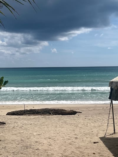A beach scene is depicted in Ban Bang Thao, Thailand, with a sandy shore in the foreground. A large piece of driftwood lies on the sand near the water's edge. The ocean features turquoise water with white waves breaking closer to the shore, and further out, the water appears a deeper blue. The sky above is a mix of dark, ominous clouds and lighter, partly cloudy areas. On the right side of the image, part of a tent or shelter is visible, supported by a pole, casting a shadow on the sand. A few leaves from a palm frond are visible in the upper left corner of the frame.