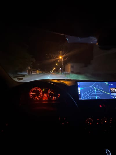 The image captures a nighttime view from inside a car driving on a road in Mogila, Bulgaria. The dashboard is visible, featuring illuminated gauges. The speedometer reads between 20 and 40 km/h. The car's navigation system displays a map with a planned route. Streetlights illuminate the road ahead, and the dark outlines of buildings and trees line the roadside. The image was likely taken during nighttime conditions.