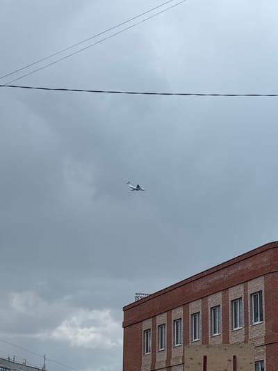 The image depicts an overcast sky with a small, white airplane flying from left to right. In the foreground, the upper portion of a red brick building with numerous windows is visible, extending from the bottom right corner upwards and towards the left. Above the building, power lines stretch across the frame. The scene is set in Dubovaya Roshcha, Russia.