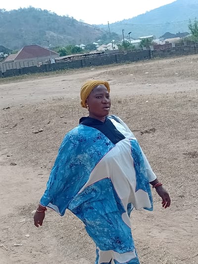 An outdoor scene in Abobo, Nigeria, depicts a woman standing on dry, light brown earth. The woman, positioned slightly left of center, wears a yellow headwrap and a loose-fitting garment that is predominantly blue with a light blue and white abstract pattern, combined with white/cream and black sections. Several red bangles are visible on her left wrist. She looks towards her right (viewer's left) with a neutral expression and slightly open mouth. The ground is sparsely covered with dry vegetation. In the background, a low concrete wall separates the foreground from residential structures, including one building with a reddish-brown tiled roof. Utility poles with overhead lighting fixtures are visible behind the wall. Distant, hazy hills or mountains rise under a bright sky in the far background.