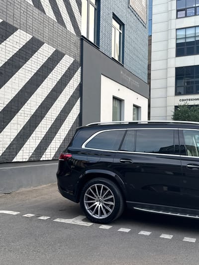 A black SUV is parked on a city street in Zhabkino, Russia. The vehicle is positioned alongside a building with a striking black and white striped facade. The building to the right is a modern office building with large windows and is labeled with Russian text. The ground is asphalt, marked with dashed white lines. The weather appears to be overcast, as indicated by the muted lighting and reflections. The image captures a typical urban scene with architectural elements and a parked car.