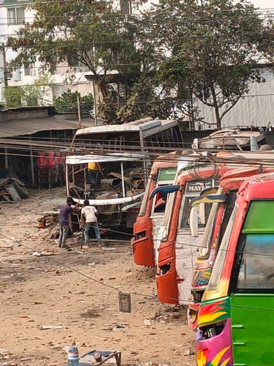 The scene in Boliarpur, Bangladesh, depicts an outdoor facility, likely a bus dismantling or repair yard. In the foreground, the right front portions of four buses are visible, painted in vibrant colors including red, green, pink, and yellow, appearing parked in a row. Labels such as "MAYER" and "Sami" are legible on some of these vehicles. The midground features a heavily damaged, primarily white bus chassis, largely stripped of its exterior panels and roof, exposing its internal structure and debris. Three individuals are present near this damaged bus: one in a yellow shirt is positioned within the bus's interior, while two others, wearing a dark purple shirt and a light-colored shirt respectively, are on the unpaved dirt ground directly in front of the bus, engaged in apparent work or dismantling activities. The ground is uneven, dusty, and littered with various metallic and non-metallic debris. Overhead and at ground level, multiple electrical cables or wires traverse the scene. In the background, trees, corrugated metal structures, and a multi-story white building with visible windows are present. A plastic water bottle rests on a low object in the lower-left foreground.