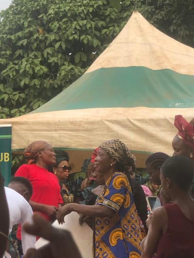 The image depicts an outdoor gathering in Awka Etiti, Nigeria, under partial natural light. A large, off-white tent with a prominent horizontal green stripe across its lower section dominates the upper mid-ground, with a dense canopy of green tree foliage visible behind it. A green banner or sign with "UB" visible is partially obscured on the far left, beneath the tent.

Multiple individuals are present, primarily women, positioned in the mid-ground and foreground. On the left, a woman wearing a red short-sleeved top and a patterned light-brown headwrap is visible from the chest up, facing right with a slight smile. Behind her, another woman wearing sunglasses and a dark headwrap is partially visible.

In the center-right, an older woman with dark skin is prominently featured, wearing a blue dress with a repeating pattern of yellow and black motifs, and a patterned dark headwrap. She is facing right and smiling, with her hands positioned as if holding a light-colored object, possibly a document or fabric.

Several other individuals are partially visible around and behind the central figures. One woman to the right of the woman in blue wears a maroon headwrap. On the far right, the back of a person, likely a younger individual, is visible holding a smartphone in their right hand, pointed towards the scene. In the immediate foreground, parts of individuals, including a person in a white shirt on the far left, are present, with hands visible, partially obscuring the lower portion of the scene.