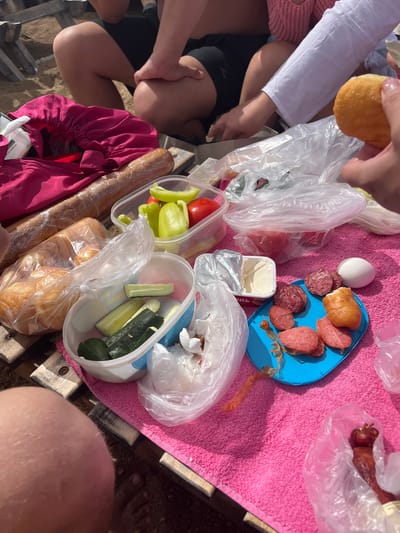 The image depicts a picnic scene with food spread out on a pink towel and wooden surfaces. Several individuals, partially visible, are present. In the foreground, a person's hand holds a piece of fried dough. Laid out are various food items including bread, sliced cucumbers in a plastic container, chopped bell peppers and tomatoes in a clear container, sliced sausages on a blue plate, a small tub of cream cheese, an egg, and a piece of fried dough. There are also plastic bags containing other food items. The setting appears to be outdoors, possibly a beach or park, with sand visible on the ground. The overall atmosphere suggests a casual gathering or meal.