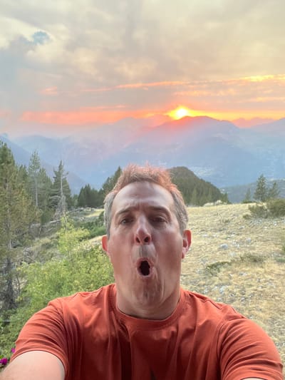 A man with an open mouth and surprised expression takes a selfie in front of a mountainous landscape at sunset. The sun is setting behind the mountains, casting a warm glow over the scene. The man is wearing an orange t-shirt. The background features a valley with trees and hills. The location appears to be a natural setting, possibly in San Juan de Plan, Spain.