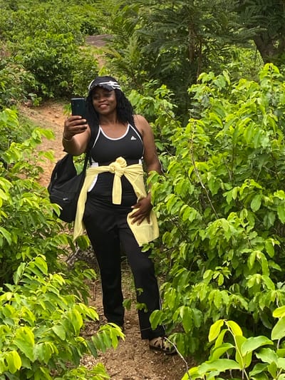 A middle-aged Black woman stands on a dirt path surrounded by lush green foliage. She is wearing a black and white tank top, black pants, and sandals. A yellow garment is tied around her waist. She is holding a smartphone in her left hand, presumably taking a selfie. A black backpack is slung over her shoulder. The location is Ganawuri, Nigeria.