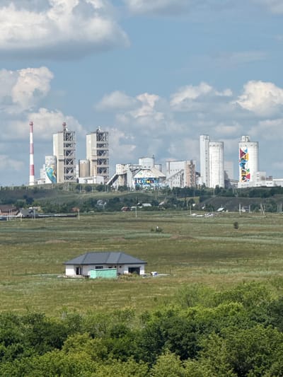 The image depicts a wide shot of an industrial complex on a hilltop overlooking a rural landscape with a single building in the foreground. The industrial complex, likely located in Novotroitsk, Russia, features several tall, white structures, including silos and towers. One prominent tower is red and white striped. The buildings have various geometric designs and some appear to be under construction or maintenance. The sky above is blue with scattered white clouds. The foreground shows a vast expanse of green grass with a modern, partially built house and a small green structure in the middle ground. Dense green trees line the bottom of the frame. The scene appears to be a clear day with bright sunlight. No people are visible in the image.