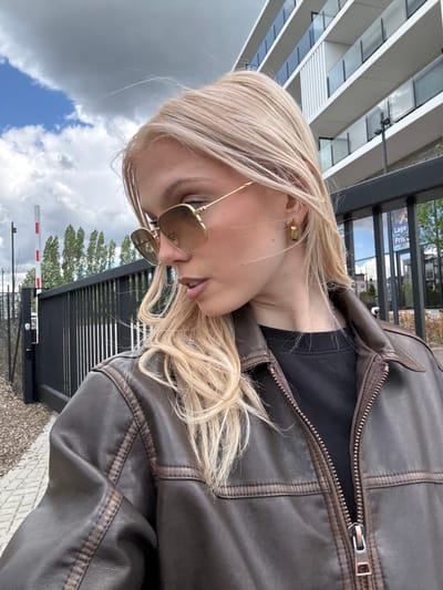 A casual, vertically oriented shot captures a young woman from the chest up in an outdoor setting in Uccle - Ukkel, Belgium. She has long, wavy blonde hair and is looking downwards and slightly to her right, wearing gold-framed sunglasses. Her attire consists of a dark brown leather jacket with visible stitching and a partially unzipped front, revealing a black top underneath. A simple gold earring adorns her left ear.
The background features a blend of urban elements and natural scenery. To the left, a modern building with multiple windows is visible. Behind the woman, there's a dark metal fence, and further back, lush green trees. A red and white automatic barrier gate, typically used for controlled access, is prominent in the mid-ground. The sky above is partly cloudy, displaying both dark grey clouds and patches of blue sky, suggesting daytime weather. On a sign visible through the fence, the partial text "Lage Prijs" (Dutch for "Low Price") can be seen. The ground beneath appears to be a paved area. The overall scene suggests a relaxed moment in a transitional space, possibly near a commercial or residential complex.