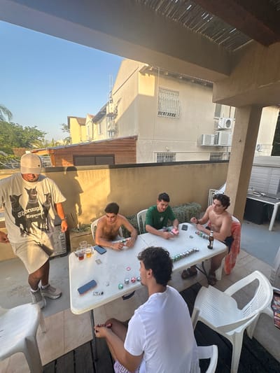 In Zoran, Israel, on a patio, four young men are gathered around a white plastic table playing a card game, likely poker, indicated by the presence of poker chips and cards. The man on the left, wearing a tan baseball cap and a t-shirt with a dog graphic, stands with his back to the camera. To his right, a shirtless man in a green t-shirt sits with his hands on the table, surrounded by poker chips. Another shirtless man, wearing dark shorts, sits opposite him and appears to be holding cards. In the foreground, a young man wearing a white t-shirt and patterned shorts sits facing away from the camera, also engaged in the game. The scene takes place outdoors, with buildings and trees visible in the background under a clear blue sky. The patio is furnished with plastic chairs and a table, and the presence of drinks suggests a casual gathering.