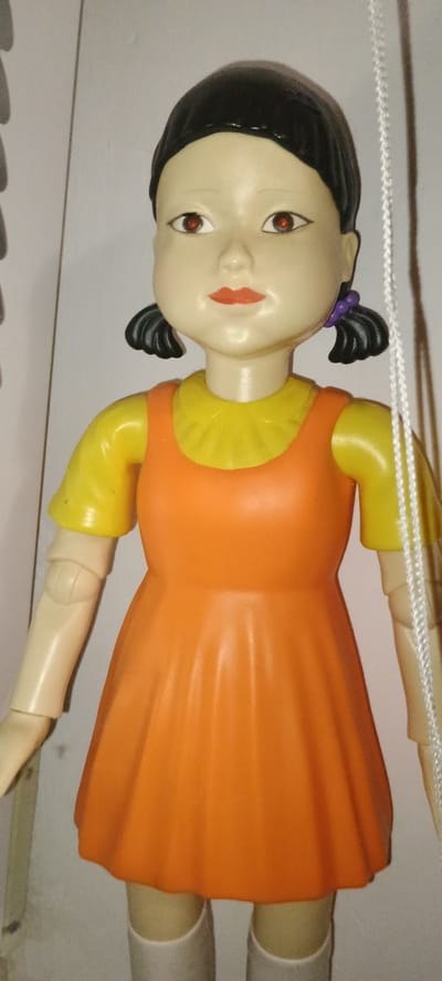 A plastic doll, resembling a character from the "Squid Game" series, is depicted against a neutral background, likely an indoor setting. The doll has a pale complexion, black hair styled in pigtails with purple ties, and bright red eyes. It is wearing a yellow shirt with a ruffled collar and an orange sleeveless dress. The doll's limbs are articulated. A white string, possibly part of a puppet mechanism, is visible on the right side of the image. The doll is positioned centrally, facing forward. The background is a pale, textured wall, with a portion of a darker, vertical element on the left side. The lighting is direct, casting soft shadows. There is no indication of the city of Pervomaisk, Ukraine, in this image; the scene appears to be a close-up of the doll itself.