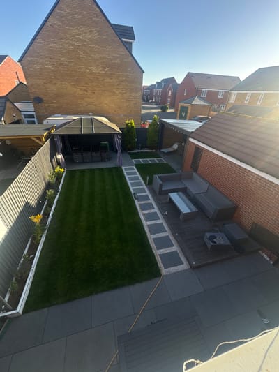This is a high-angle, eye-level, exterior shot of a modern backyard in Peterborough, United Kingdom. The yard is divided into several distinct areas. A neatly manicured lawn occupies the left-central portion of the image, bordered by a grey metal fence on the left and a pathway of square grey tiles interspersed with white gravel on the right. Adjacent to the lawn, under a hexagonal gazebo with a dark grey canopy and curtains, is a seating area with four dark grey woven storage units. To the right of the lawn and gazebo, a wooden deck hosts a grey woven L-shaped sofa and a glass-topped coffee table. Further to the right, a charcoal-colored fire pit sits next to two dark grey woven cube seats. The right side of the yard features a red brick house with a brown tiled roof. The entire scene is illuminated by bright sunlight, casting distinct shadows. In the background, rows of modern brick houses with tiled roofs are visible under a clear blue sky.