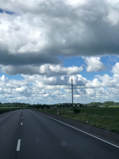 The image depicts a view from a moving vehicle on a paved, two-lane road in Dyatlitsy, Russia. The road has a solid white line on the right edge and dashed white lines marking the center. On either side of the road are green fields with scattered wildflowers and trees. In the distance, a line of trees forms a border along the horizon, with several buildings and structures visible amongst them. A tall utility pole with multiple wires is prominent on the right side of the road. The sky is filled with dramatic, heavy clouds, with patches of blue sky visible. Two cars are seen in the distance on the right side of the road.