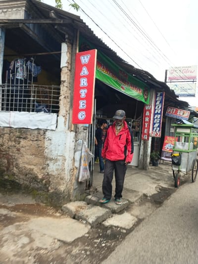 An elderly man in a red jacket and cap stands on the concrete steps leading into a modest shop front. The shop features red "WARTEG" signs, one vertical and one horizontal, and a green awning above. To the right, a food cart with a sign stands parked. Signage for "KINNAR PRATAMA JGLOW," "PHARM INDO APOTEX," and "PET SHOP" are visible above the shops. A figure stands in the shop's entrance, and clothes hang in a caged area to the left. Power lines stretch across the overcast sky.