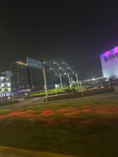 A nighttime cityscape features a high-rise building illuminated with multiple lights. Above it, a green highway sign is partially visible. Adjacent to the road, there's a well-maintained green median strip. Further along, multiple light poles are arranged. A large billboard with the word "Live" in colorful lettering is positioned at the top of the frame. The sky is dark and blurry, suggesting a moving camera.