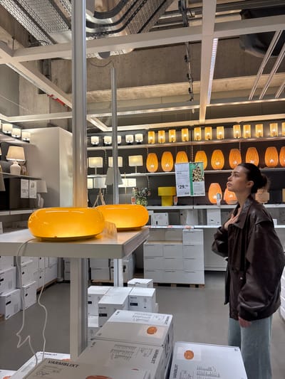 The image depicts the interior of an IKEA store's lighting section in Corsico, Italy. In the mid-ground, a female individual stands in profile, facing right and looking upwards. She wears a dark brown oversized leather jacket over light-colored pants and has dark hair pulled back. Her right hand is positioned on her chest.

In the left foreground, two glowing, oblong, orange-yellow table lamps are displayed on a white shelf. White power cords descend from these lamps. Directly beneath this display, and extending into the lower foreground, multiple white product boxes are stacked, some explicitly labeled "VÄRMBLIXT." Further back, additional white boxes marked "STOCKHOLM 2023" and "ÅRSTID" are stacked on pallets.

The background features multiple rows of various lighting fixtures displayed on wall-mounted shelves. These include white table lamps with fabric shades, small white pendant lights, and numerous orange-yellow, basket-weave-style lamps. A price tag indicating "$29.95" is visible in this area, along with a yellow rectangular display block. The ceiling reveals exposed concrete beams, white structural supports, metal cable trays, and linear fluorescent lighting fixtures. A tall, thin white pole extends vertically from the floor to the ceiling, positioned slightly left of the image center. The overall environment is brightly illuminated by artificial light.