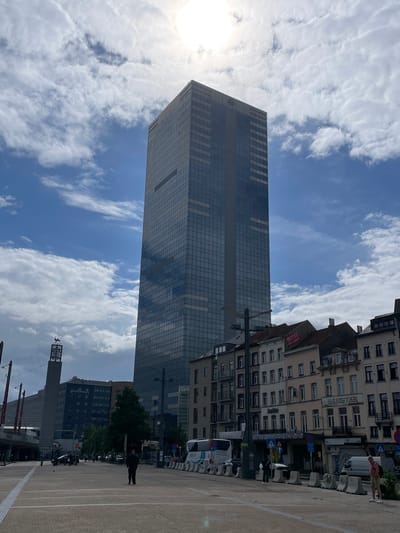 The image presents a wide-angle view of an urban landscape in Saint-Gilles, Brussels, Belgium, near the Gare du Midi. A tall, rectangular skyscraper, identified as the Tour du Midi (South Tower), dominates the central-right of the frame, clad in reflective blue-tinted glass. The upper-right quadrant of the sky is intensely bright from the sun, which is partially obscured by bright white and grey clouds scattered across an otherwise blue sky.
In the mid-ground on the left, a distinctive concrete tower with a lattice-like top supports a dark, abstract sculpture of a bird with outstretched wings, known as 'L'Envol' by Olivier Strebelle. Behind this, the "Pullman Hotel" building is visible, with dark cladding and white lettering. Elevated railway infrastructure is partially visible further left in the background.
The foreground consists of a broad, paved plaza with light-colored square paving stones and white painted lines. Grey concrete barriers separate the plaza from a parallel street on the right. A white city bus is parked on this street, along with a white delivery van. Several individuals are present: one person in dark clothing stands centrally in the plaza, facing away from the camera; another person in a light-colored shirt and shorts stands on the far right near the concrete barriers, holding a bag; and a third person with a white backpack is partially visible near the barriers. Other pedestrians are discernible in the background.
The right side of the image features a row of older, multi-story buildings with varied facades, balconies, and traditional windows. Signs include a red "HOTEL" sign on a rooftop, a "HOTEL OHFADISTA" sign below, and a "Jupiler" sign. Traffic signs, including a blue circular mandatory direction sign and a red circular no-entry sign, are visible on poles. The scene suggests a daytime setting with moderate activity.