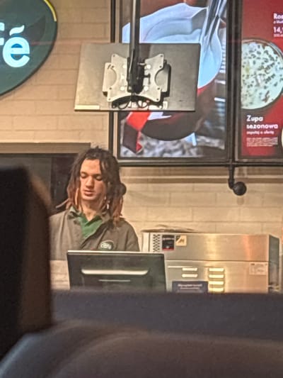 The media file shows a person with dreadlocks wearing a dark grey uniform with a green collar and a small, round logo on the left chest that says "Cafe". The person appears to be a barista or cashier. They are looking down, possibly at a screen or counter, with a neutral or tired expression. The scene appears to be inside a cafe or fast-food establishment, indicated by the presence of a stainless steel food service machine, a digital menu board, and a counter. The menu board displays Polish text and images of food items. One visible text reads "Zupa sezonowa" which translates to "Seasonal soup". The lighting suggests it is daytime, but the interior setting makes it difficult to determine the exact time. The background shows tiled walls and a dark, blurred foreground which might be another customer or furniture. The person's posture and the overall ambiance suggest a mundane work environment.