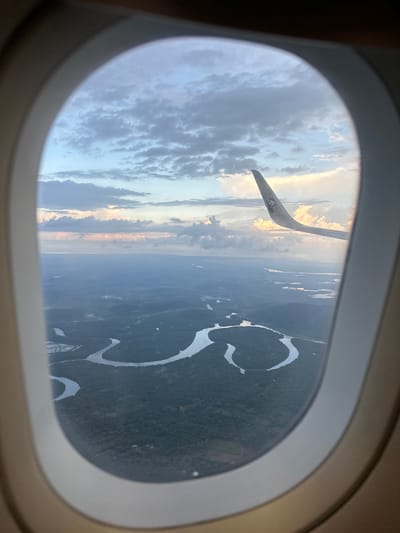 The image, taken from an airplane window, offers a panoramic view of a lush, green landscape characterized by meandering rivers and dense forestation. The sky above is adorned with scattered clouds, illuminated by the soft light of either dawn or dusk, casting a serene and slightly hazy glow over the scene. A portion of the airplane's wing is visible on the right side of the frame, extending towards the clouds. While the precise location within Ayer Tawar, Malaysia is not discernible from the image itself, the topography suggests a region with significant water bodies and extensive natural vegetation, typical of many areas in Malaysia. There are no people or specific human activities directly visible in the image, apart from the implied presence of the aircraft and its passengers. The overall impression is one of natural beauty viewed from a high vantage point.